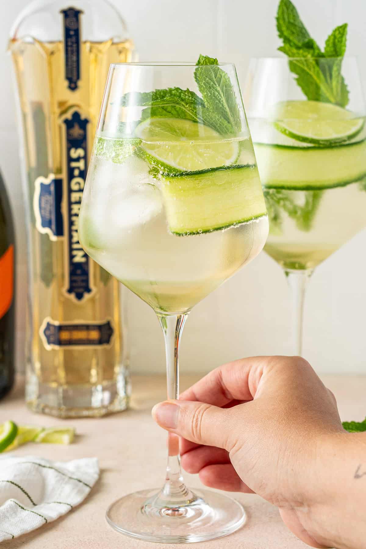 A hand holding the stem of a glass of a Hugo spritz cocktail. 