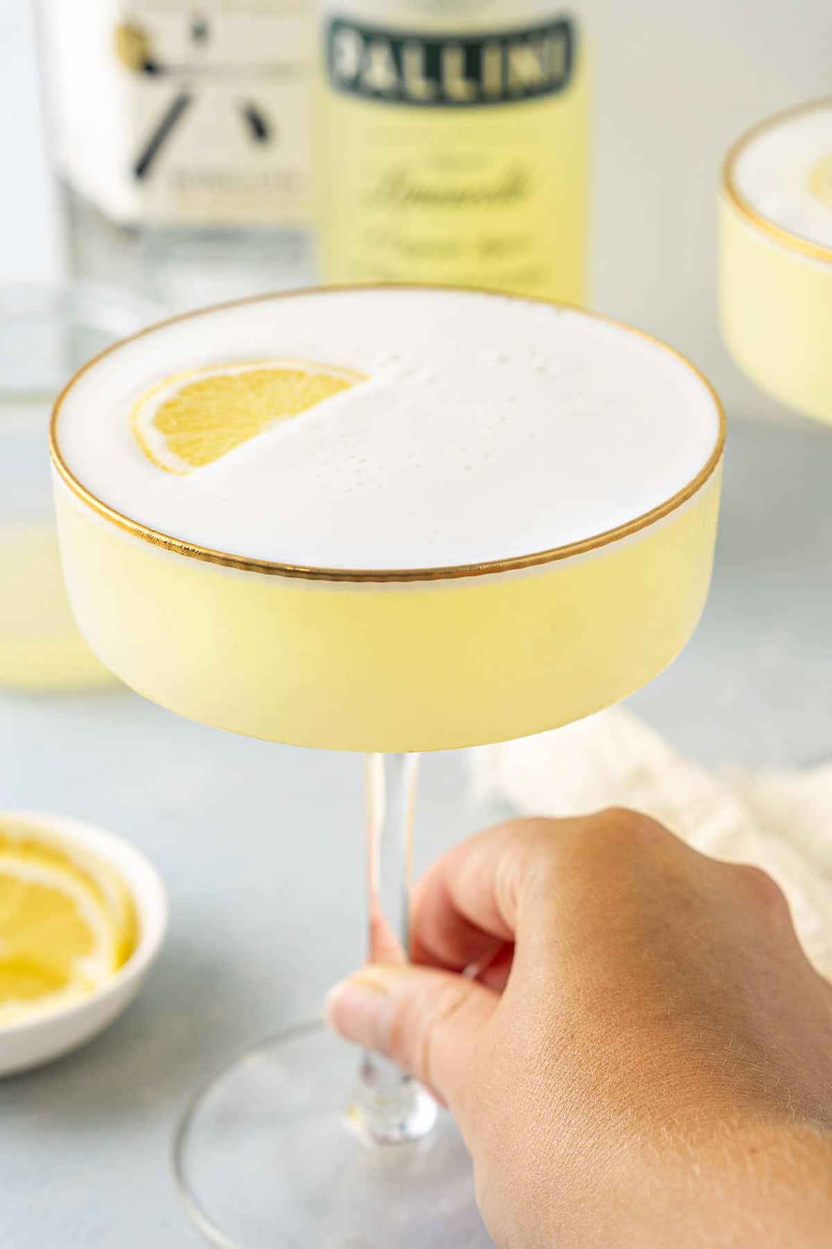 A hand holding the stem of a limoncello sour in a glass.