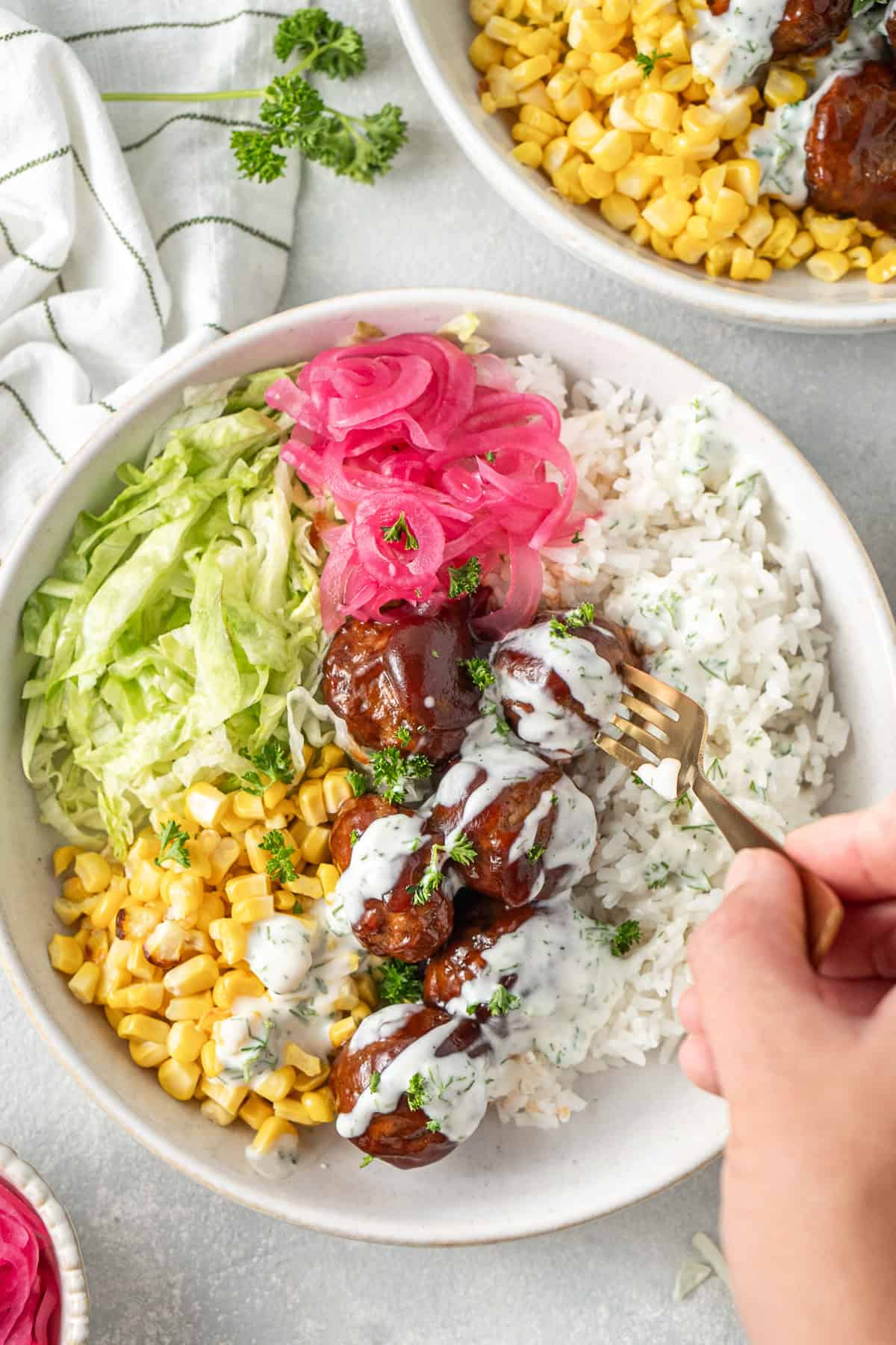 A hand holding a fork with the BBQ beef and rice bowl with pickled onions and corn.