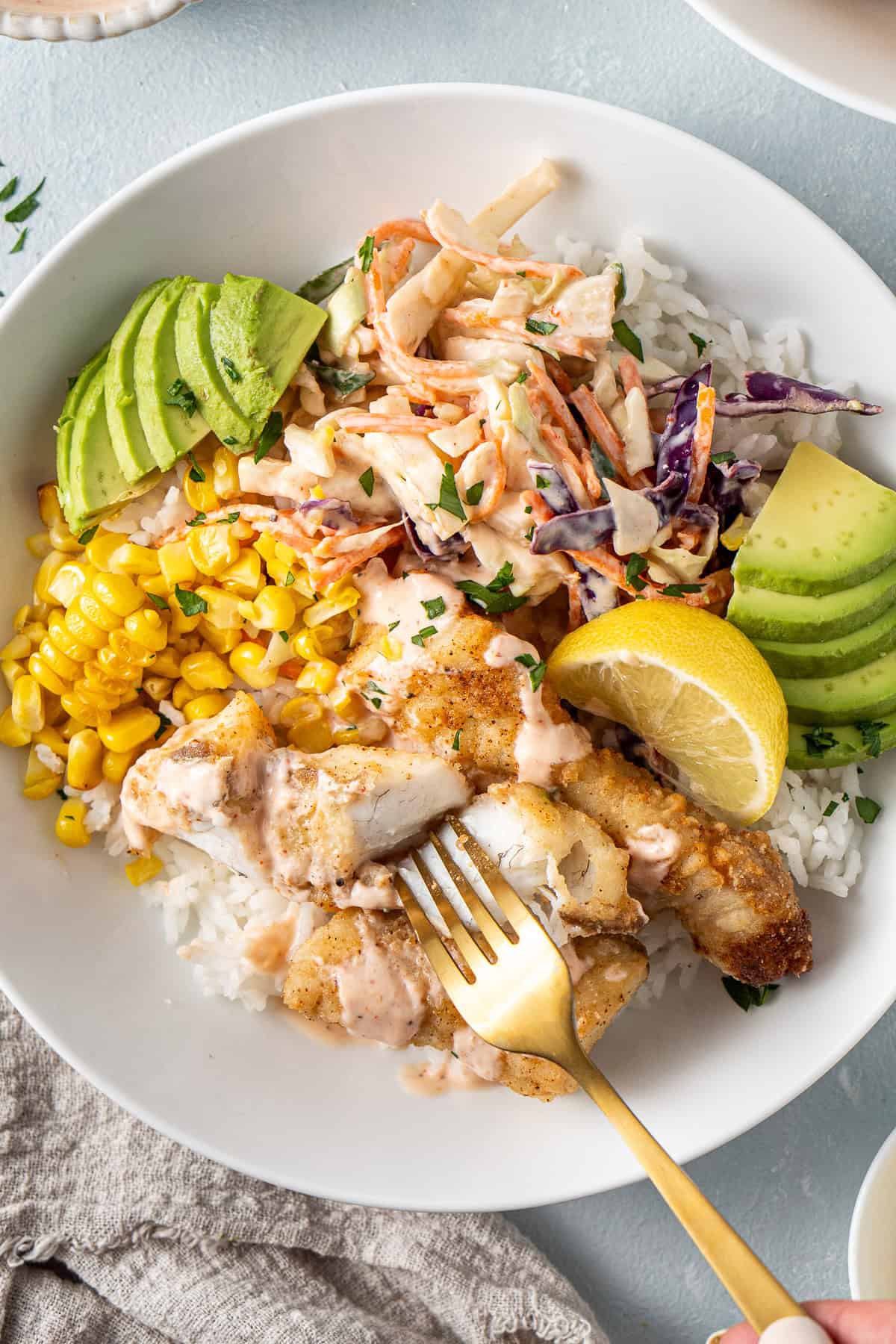 A fork breaking into the white fish flesh in a taco bowl with fresh lemon and slaw. 