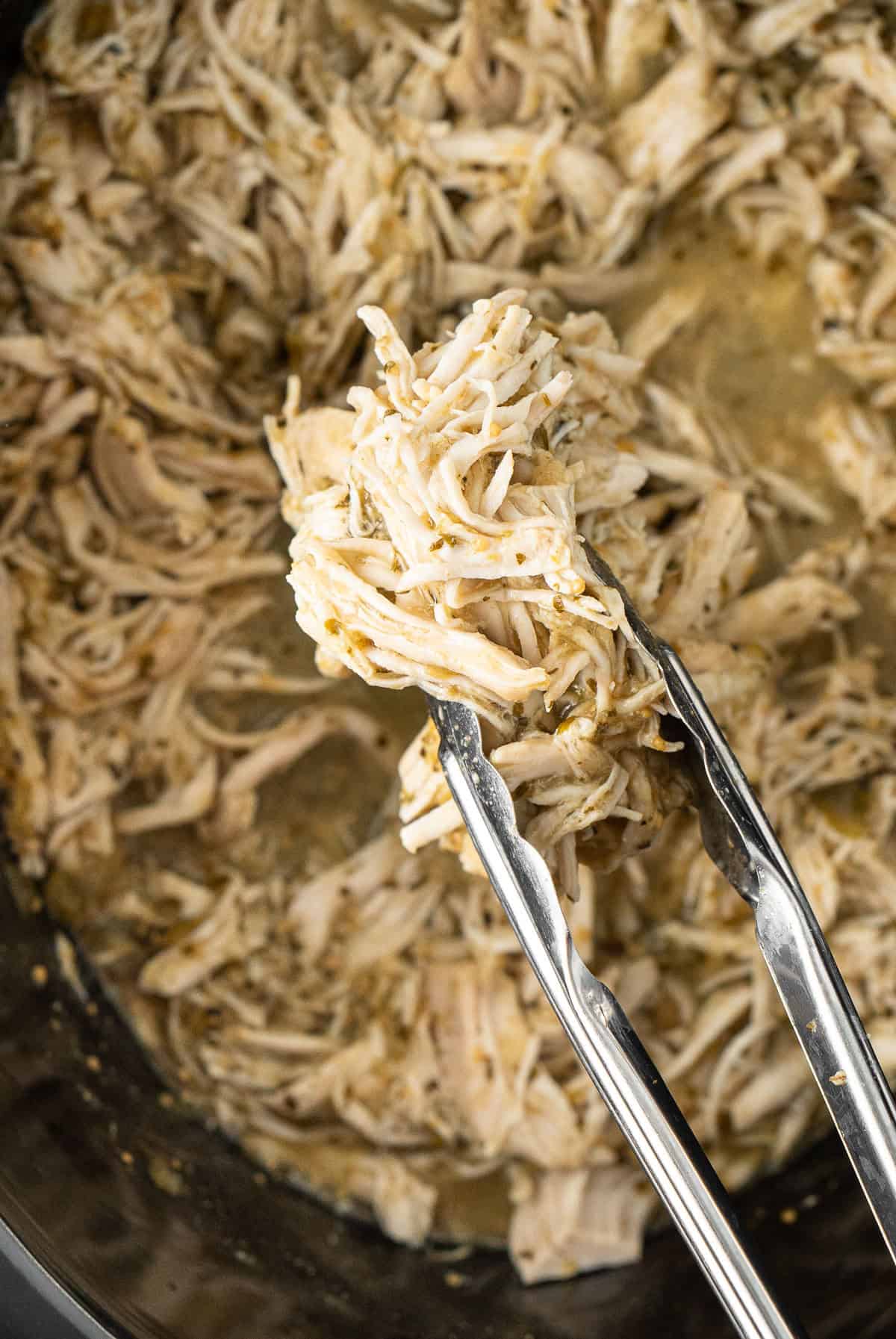 Tongs holding the shredded chicken putting it back in the slow cooker.