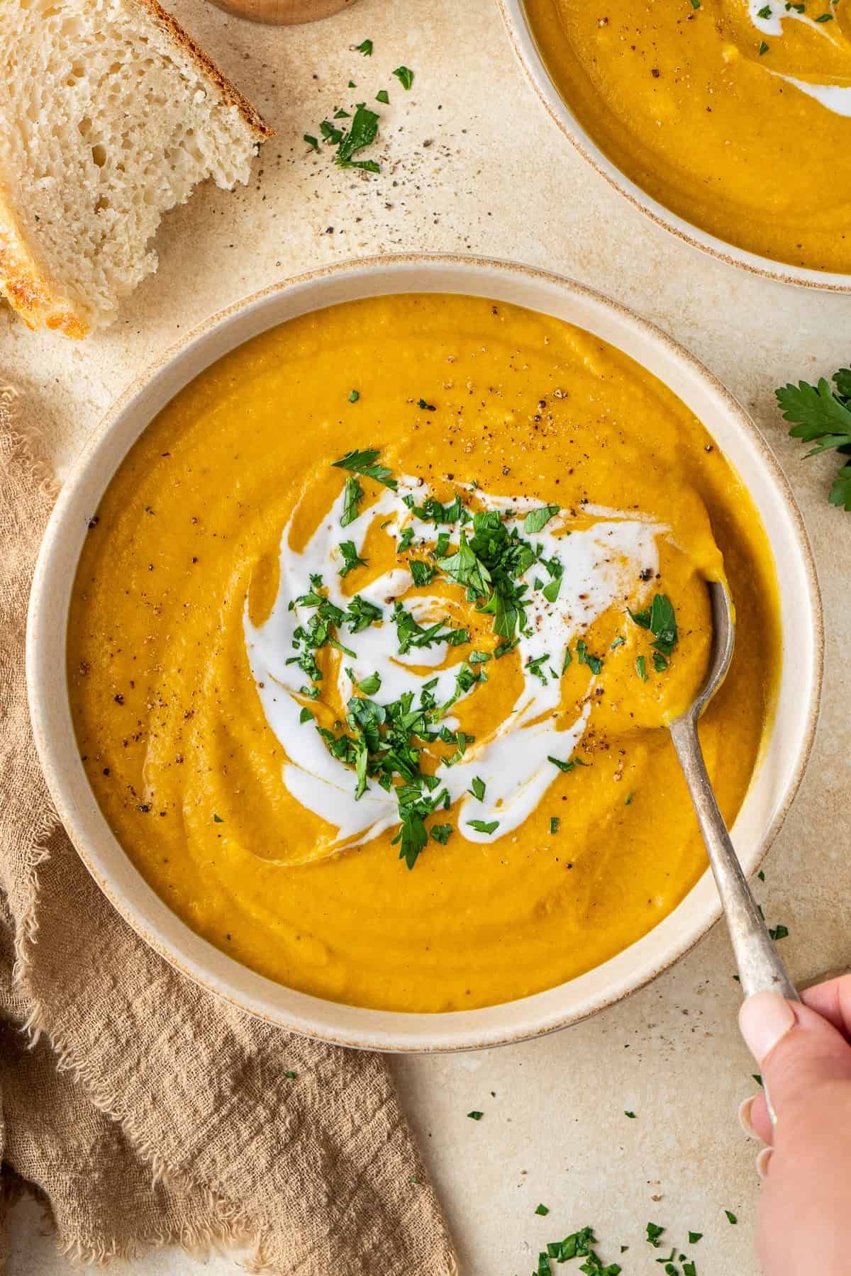 A hand holding a spoon taking a scoop of carrot lentil soup topped with coconut milk and chopped parsley.