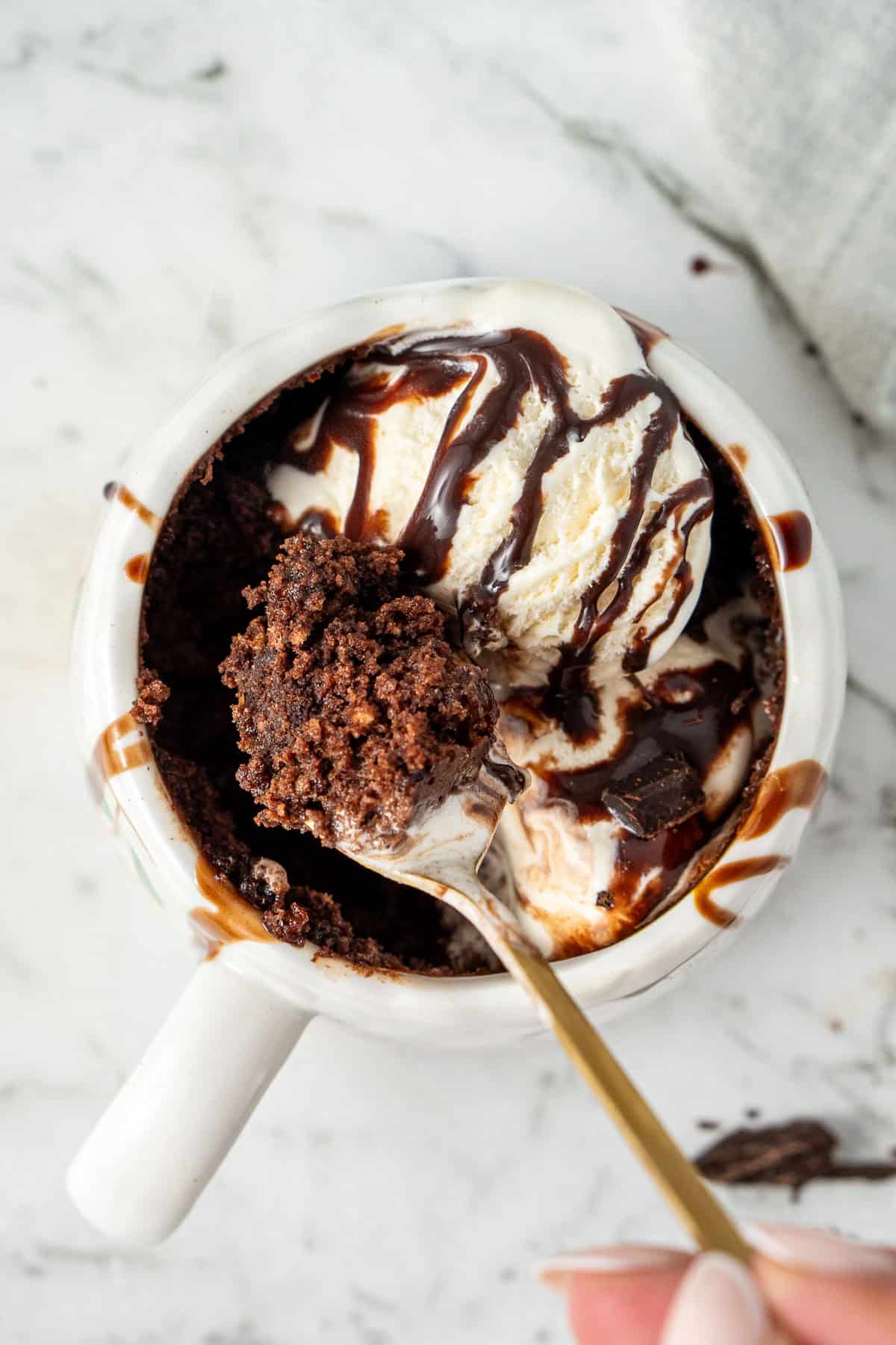 A spoon taking a bite of chocolate mug cake with ice cream and chocolate sauce.