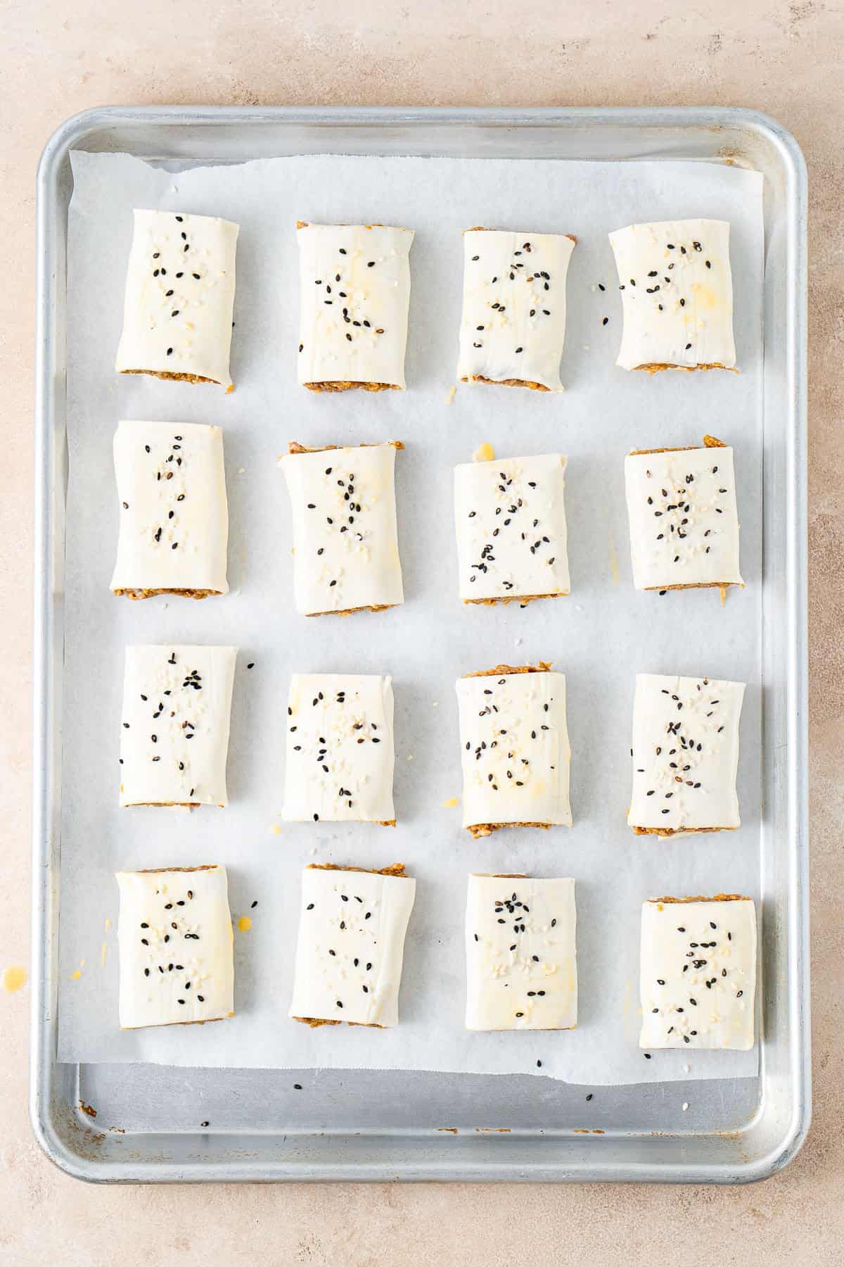 The uncooked sausage rolls on a baking tray with sesame seeds.