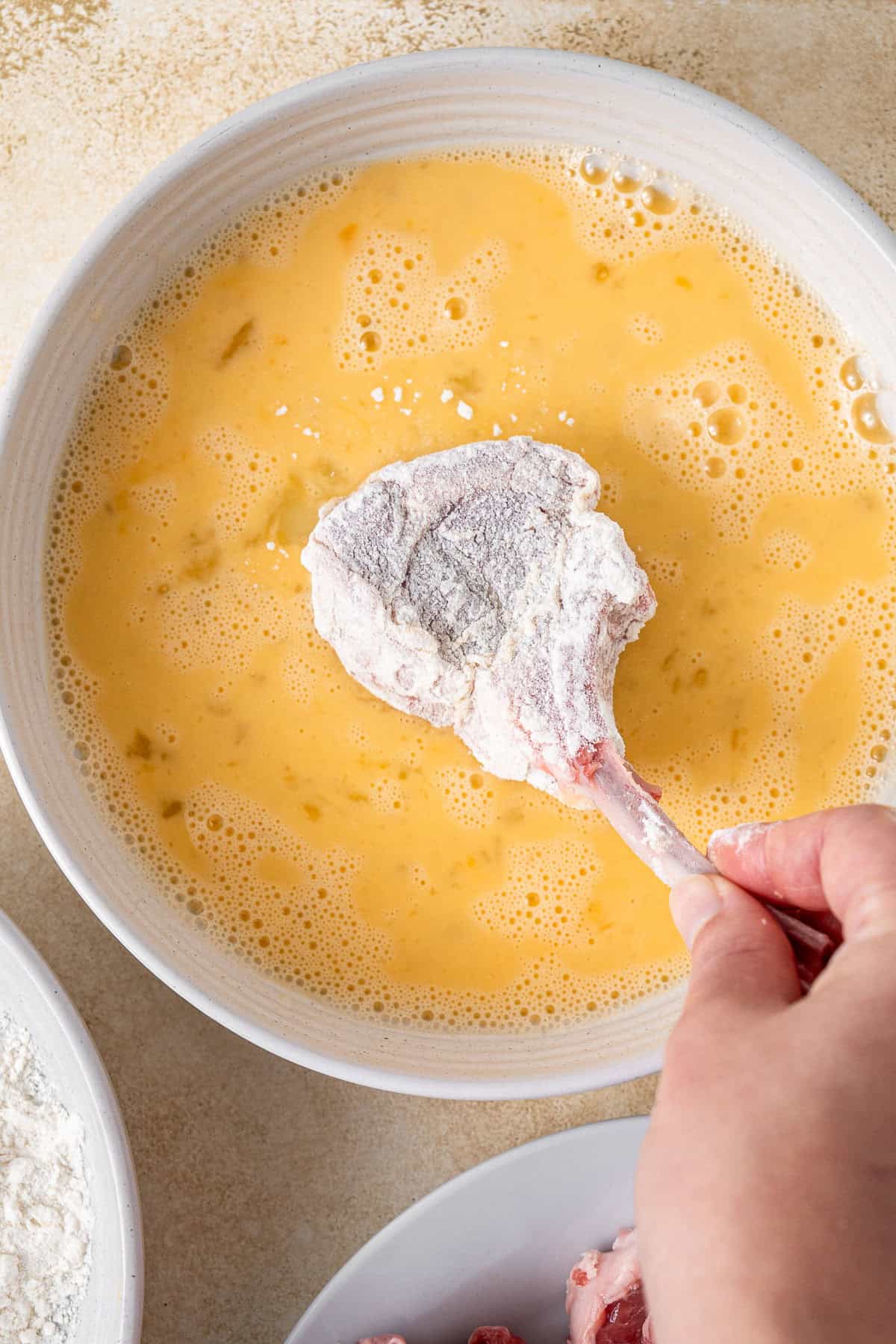 A cutlet coated in flour being dipped in egg.
